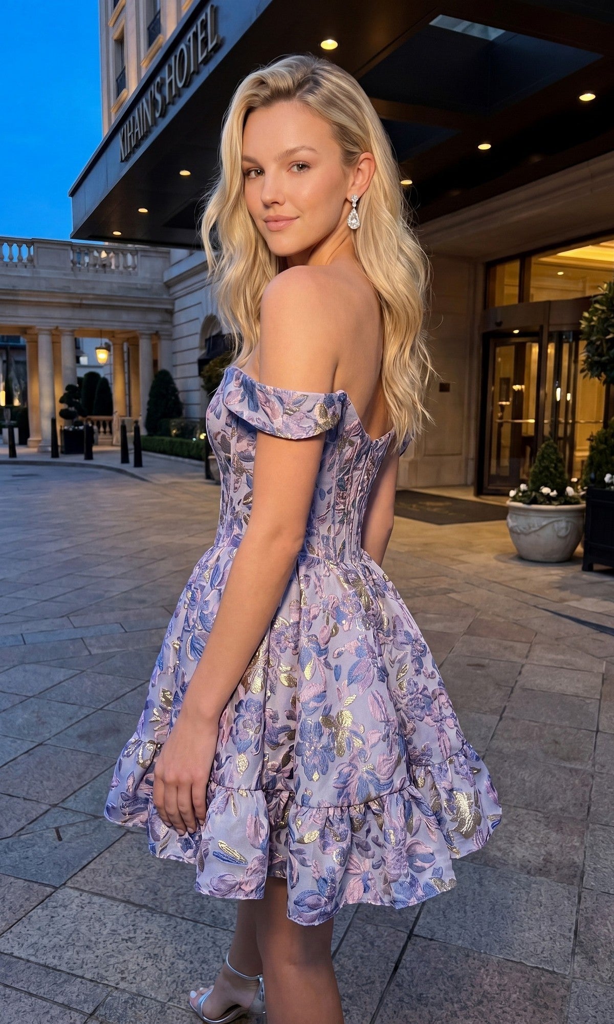A woman wearing the Nox Anabel A7018 short homecoming dress stands outside a hotel entrance in the evening, glancing over her shoulder at the camera.